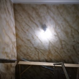 Usługi Elektryczno Budowlane Leszek Drewczyński - Ściana z efektem dekoracyjnym w odcieniach beżu i złota, oświetlona lampą. Widoczne fragmenty rusztowania i poręczy. Dekoracyjne malowanie ścian.