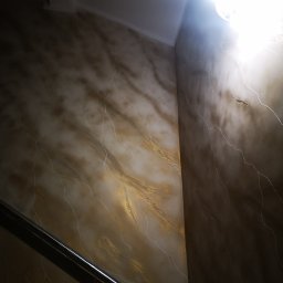 Usługi Elektryczno Budowlane Leszek Drewczyński - Dekoracyjna ściana w odcieniach brązu i złota z efektem marmuru, oświetlona lampą sufitową. Widoczny fragment metalowej listwy przypodłogowej.