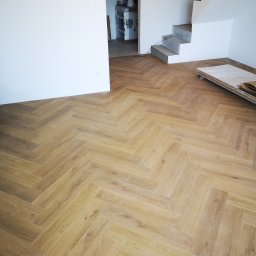 Usługi Elektryczno Budowlane Leszek Drewczyński - Ułożona podłoga z paneli w jodełkę, jasne drewno, białe ściany w tle, surowe schody betonowe, częściowo widoczne materiały budowlane.