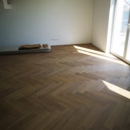 Usługi Elektryczno Budowlane Leszek Drewczyński - Ułożona parkietowa podłoga w jodełkę w jasnym, pustym pokoju z dużym oknem i stertą paneli pod ścianą. Widoczne promienie słoneczne padające na podłogę.