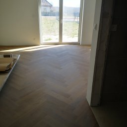 Usługi Elektryczno Budowlane Leszek Drewczyński - Ułożona parkietowa podłoga w jodełkę w jasnym wnętrzu z dużym oknem. Częściowo widoczne deski do montażu. Fragment surowej ściany w tle.