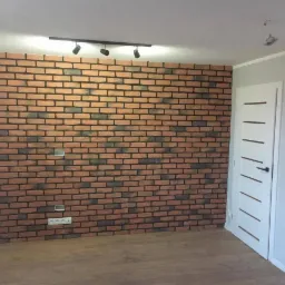 Ściana wykończona cegłą klinkierową z widocznymi puszkami elektrycznymi, oświetlenie sufitowe punktowe oraz białe drzwi z poziomymi frezami.