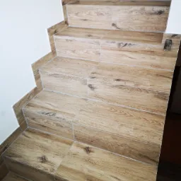 Schody wyłożone płytkami imitującymi drewno, z widocznymi fugami i narożnikami, prowadzące do góry wzdłuż białej ściany.