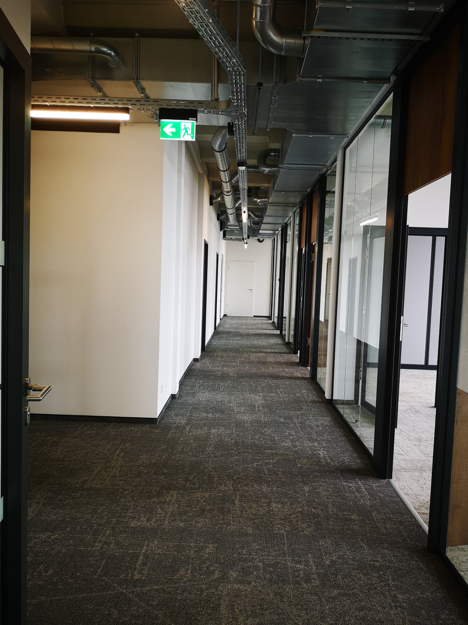 Długi korytarz biurowy z szarą wykładziną, przeszklonymi ścianami i widocznymi instalacjami pod sufitem. Zielony znak ewakuacyjny. Perspektywa z głębi korytarza.