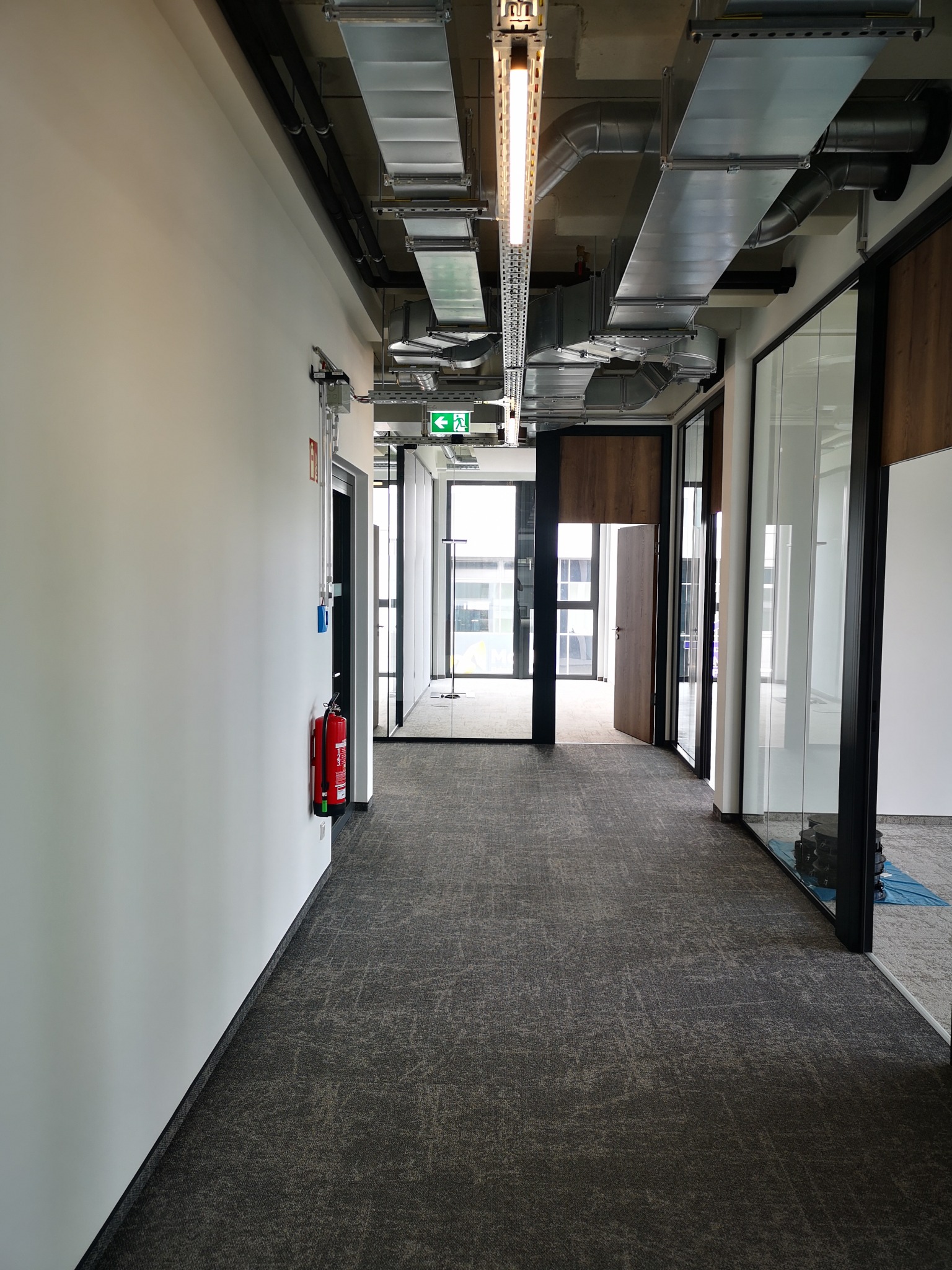 Korytarz biurowy z widocznymi instalacjami pod sufitem, ścianami pomalowanymi na biało i wykładziną podłogową w odcieniach szarości. Widoczny system wentylacji i oświetlenie.