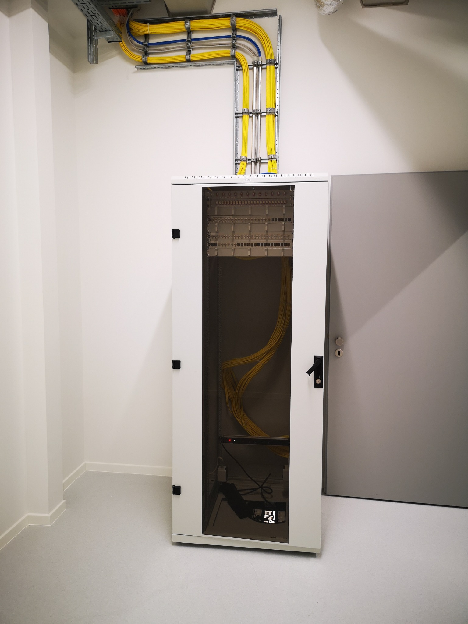 Biała szafa rack z żółtymi kablami wewnątrz, widoczne kable w korytach na suficie, szare drzwi obok. Minimalistyczne wnętrze, jasne oświetlenie.