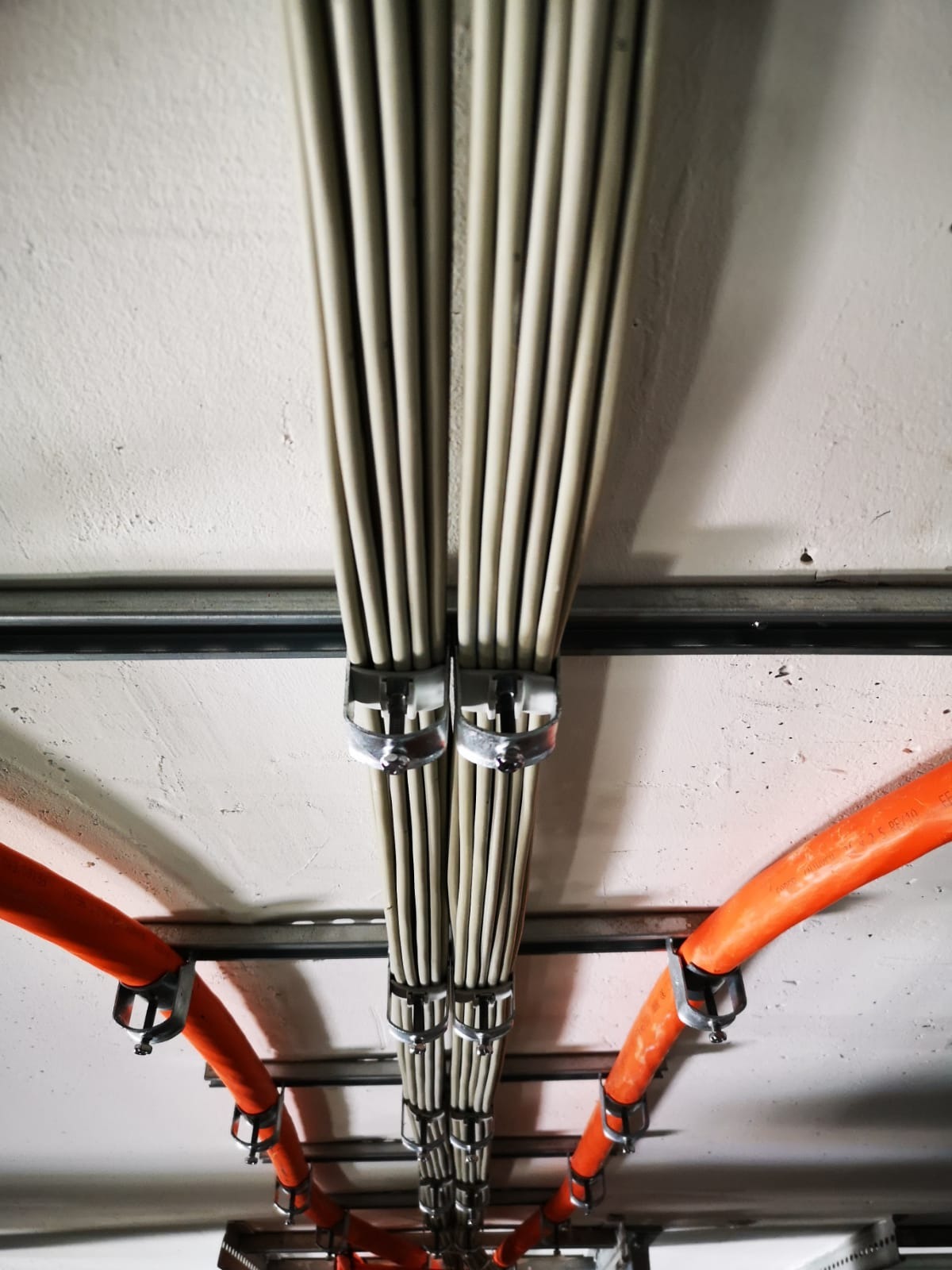 Zamocowane kable elektryczne w peszlach i wiązkach, prowadzone pod sufitem. Widoczne metalowe uchwyty mocujące i fragmenty instalacji.