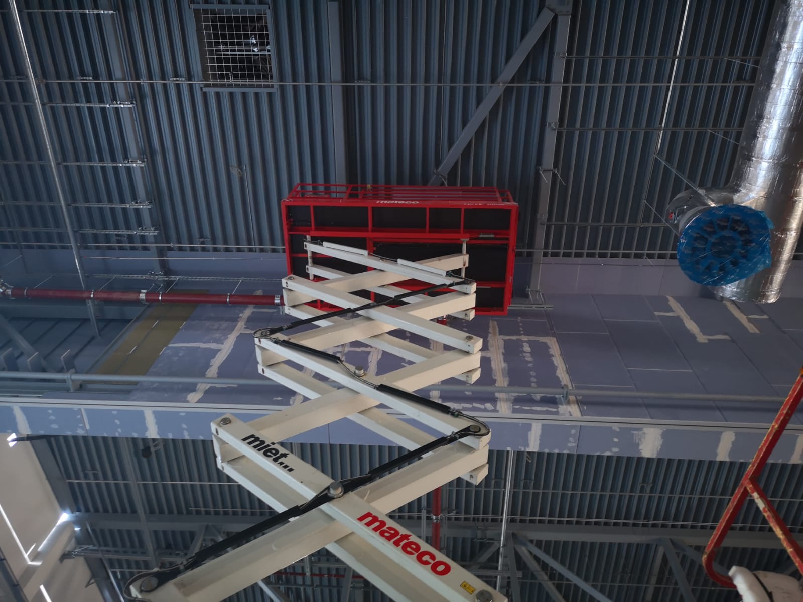 Wnętrze w trakcie wykańczania: podwieszany sufit z instalacjami, czerwony podnośnik nożycowy Mateco, widoczne płyty karton-gips i rury wentylacyjne.