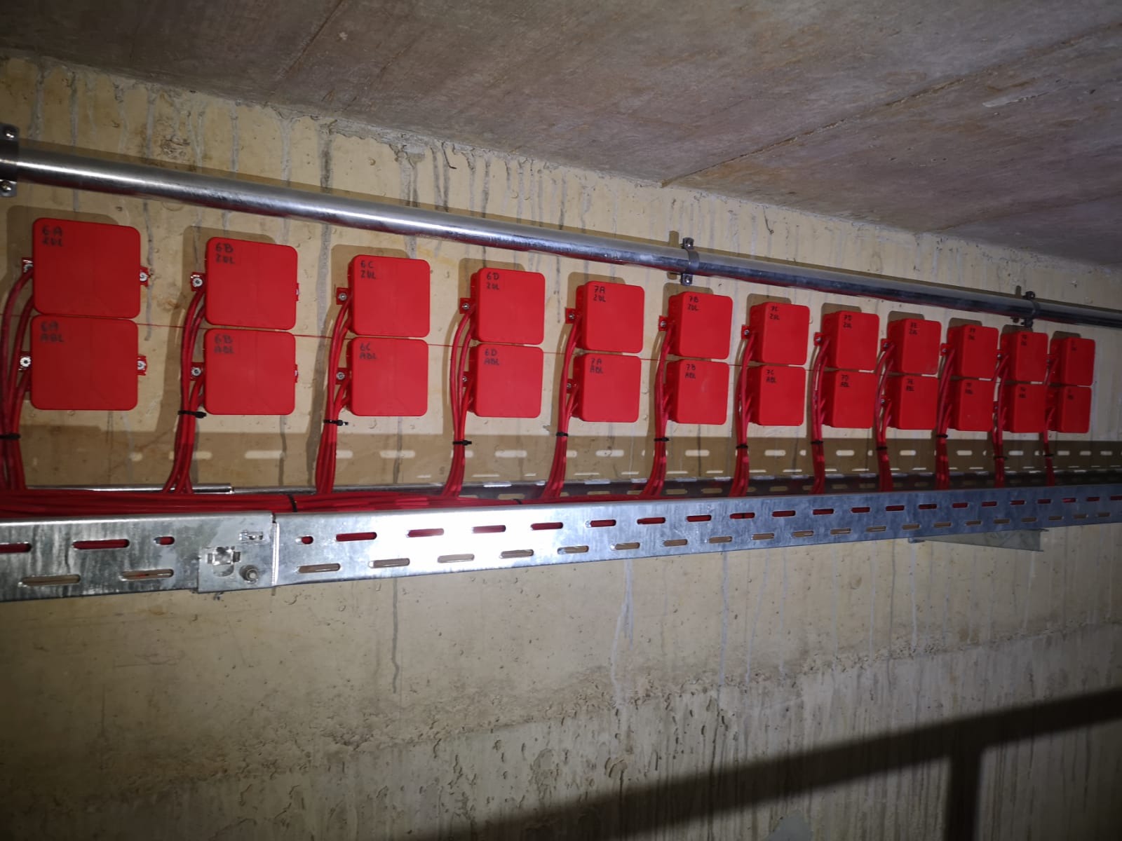 Rząd czerwonych puszek z kablami, oznaczonych numerami, przymocowanych do ściany i metalowej szyny. Detal instalacji elektrycznej, widok z dołu, surowy beton w tle.