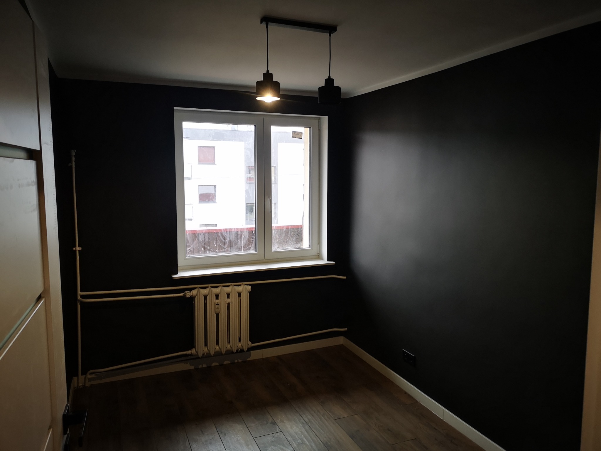 Pokój po malowaniu ścian na ciemny kolor, z drewnianą podłogą i białym oknem z grzejnikiem. Minimalistyczne lampy wiszące.