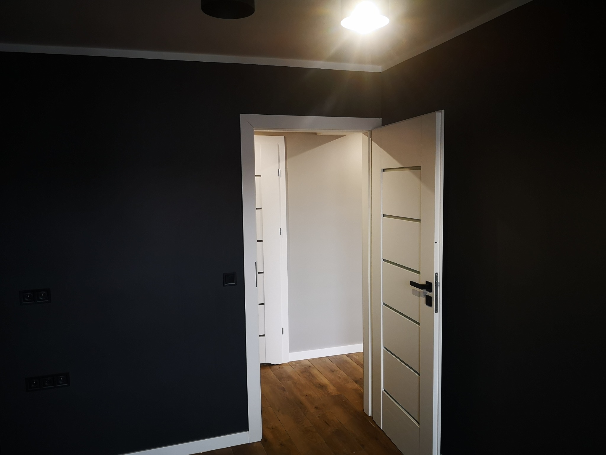 Nowoczesne wnętrze po remoncie: grafitowa ściana z gniazdkami, drewniana podłoga i białe drzwi z czarnymi klamkami, widok na sąsiednie pomieszczenie.