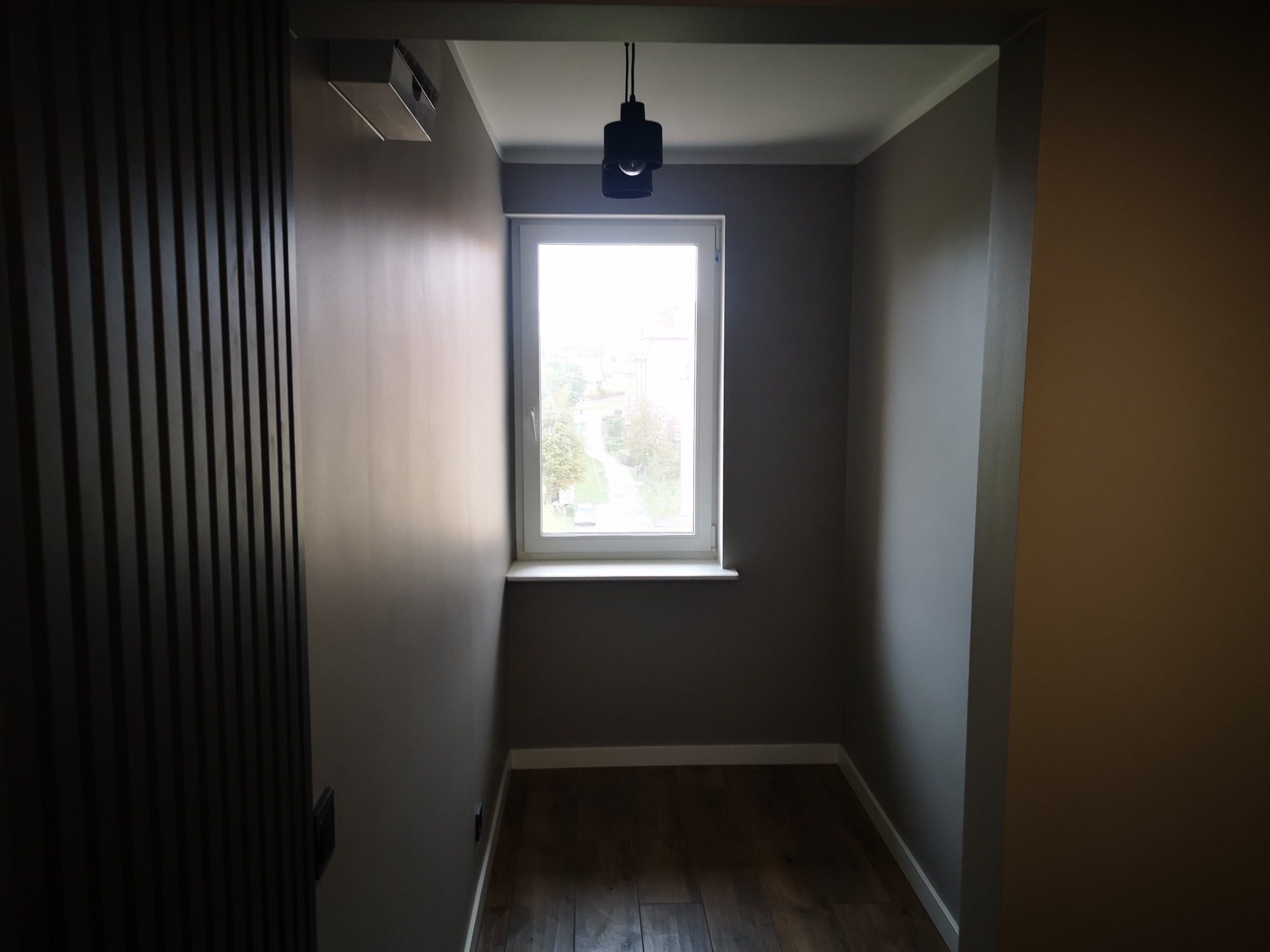 Wnętrze pokoju po remoncie: szare ściany, drewniana podłoga, okno z widokiem na zieleń, czarna lampa sufitowa i dekoracyjna ściana z drewnianych listew.