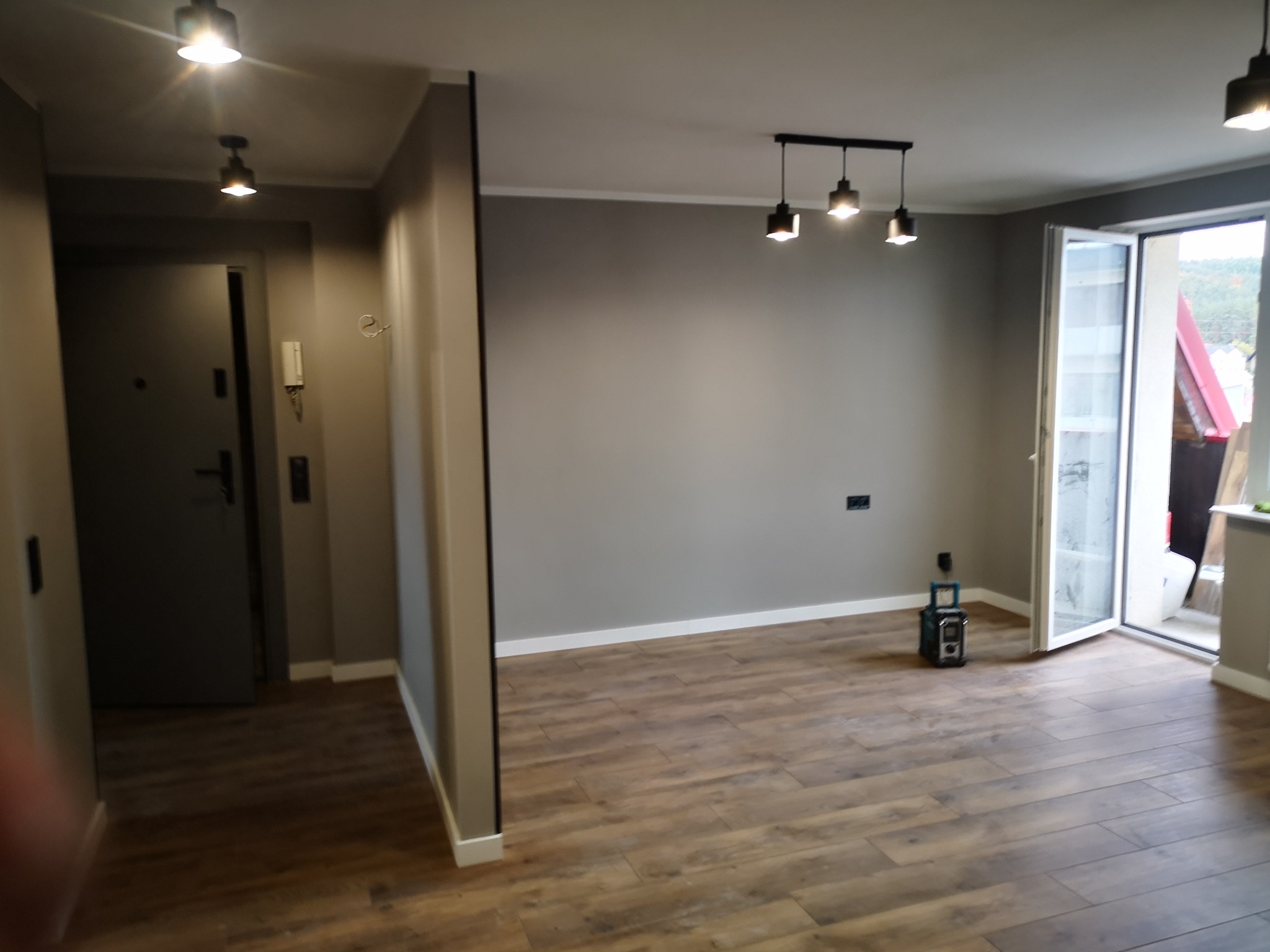 Szare wnętrze po remoncie: drewnopodobna podłoga, ściany w odcieniach szarości, nowoczesne oświetlenie sufitowe i otwarte drzwi balkonowe wpuszczające światło.