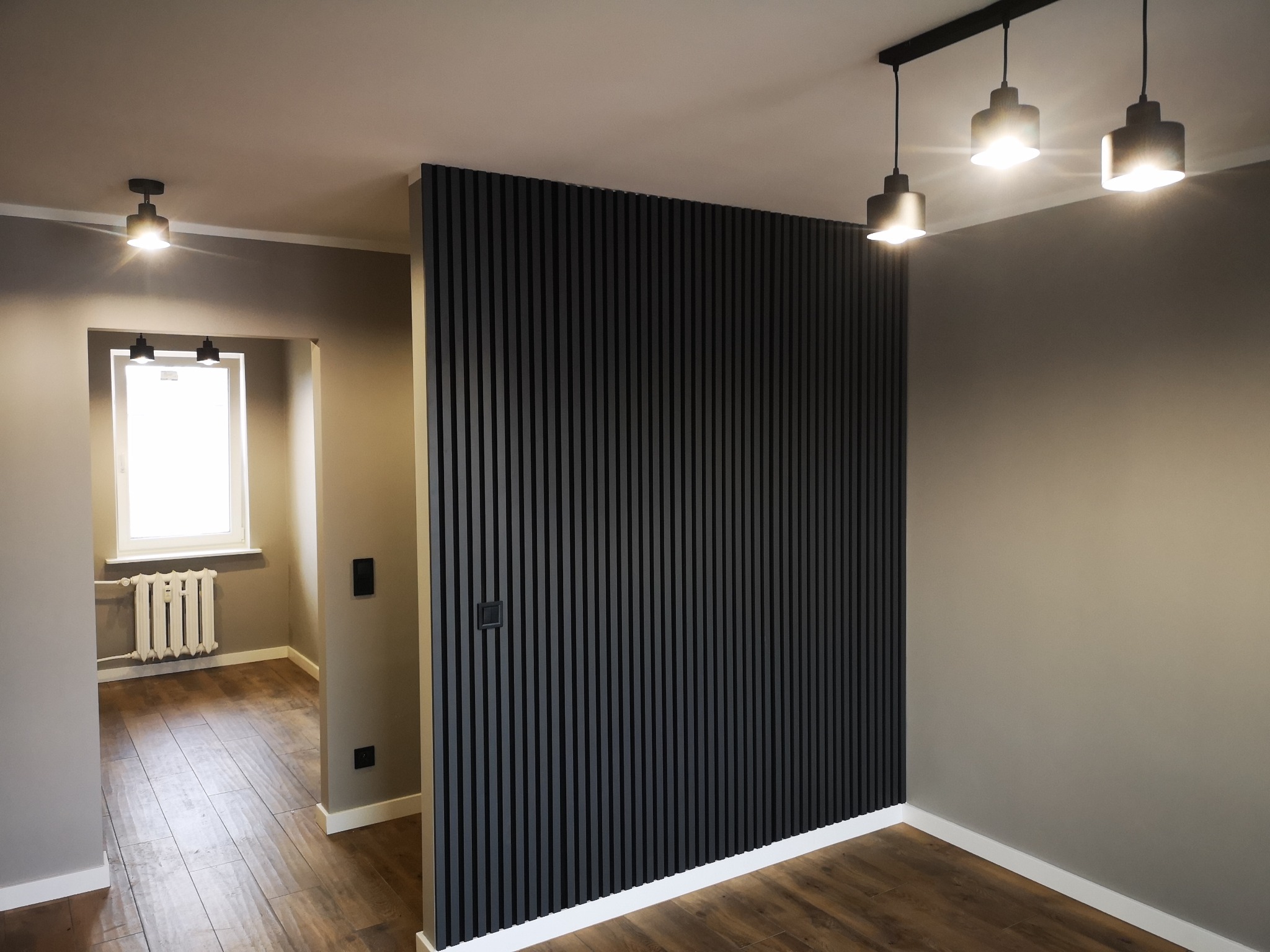 Nowoczesne wnętrze z drewnianą ścianką działową z lameli w kolorze grafitowym, widok na pokój z grzejnikiem i drewnianą podłogą, oświetlenie punktowe.