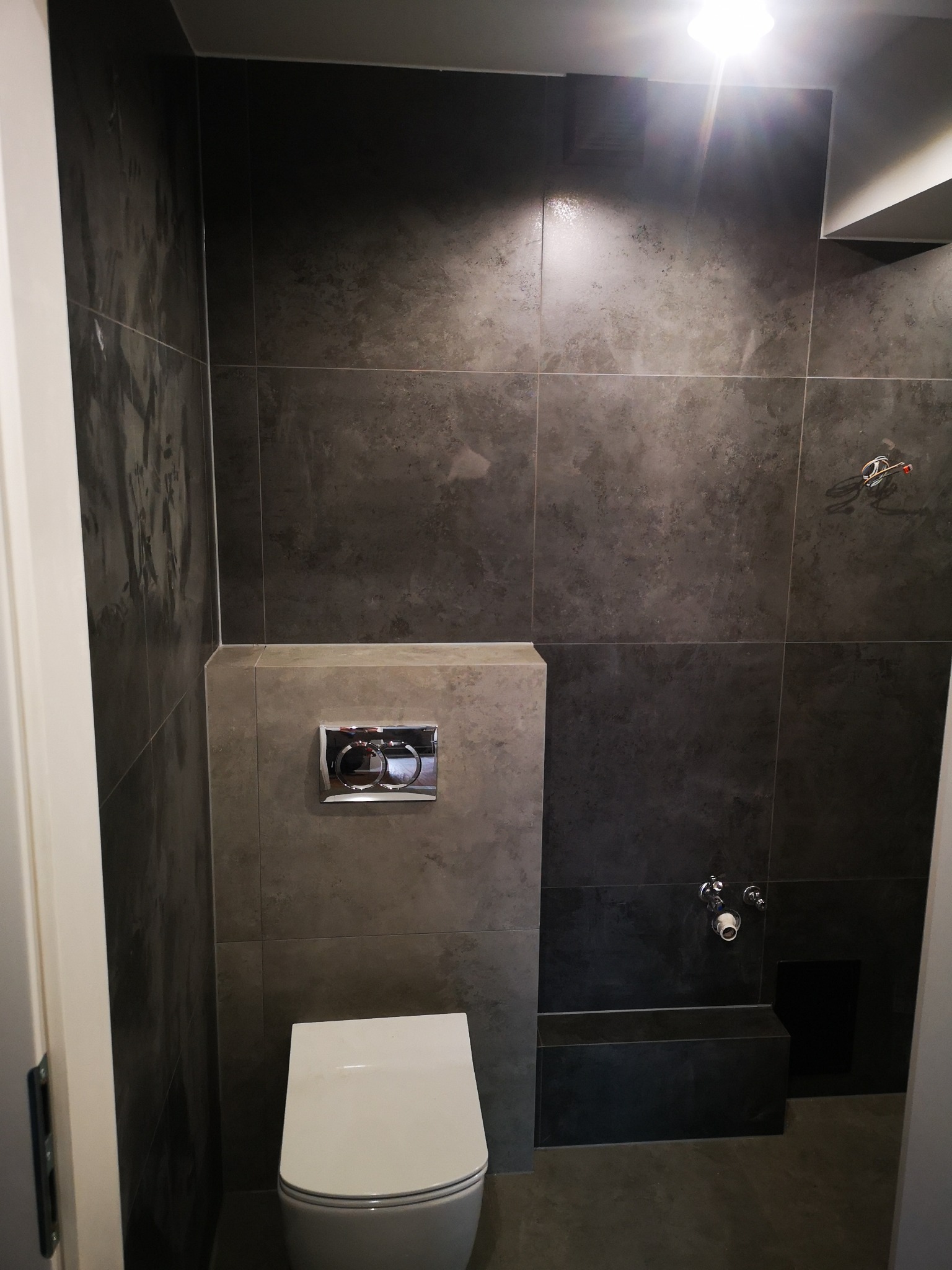 Nowoczesna łazienka z ciemnymi, grafitowymi płytkami, wiszącą toaletą i chromowanym przyciskiem spłukiwania. Widoczne przyłącza wodne i fragment instalacji elektrycznej.