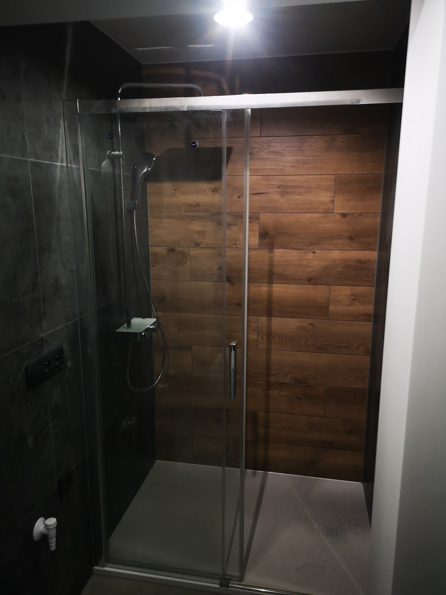 Nowoczesna kabina prysznicowa z drewnopodobnymi płytkami na ścianie, szklane drzwi, czarne płytki po lewej stronie, minimalistyczny design.