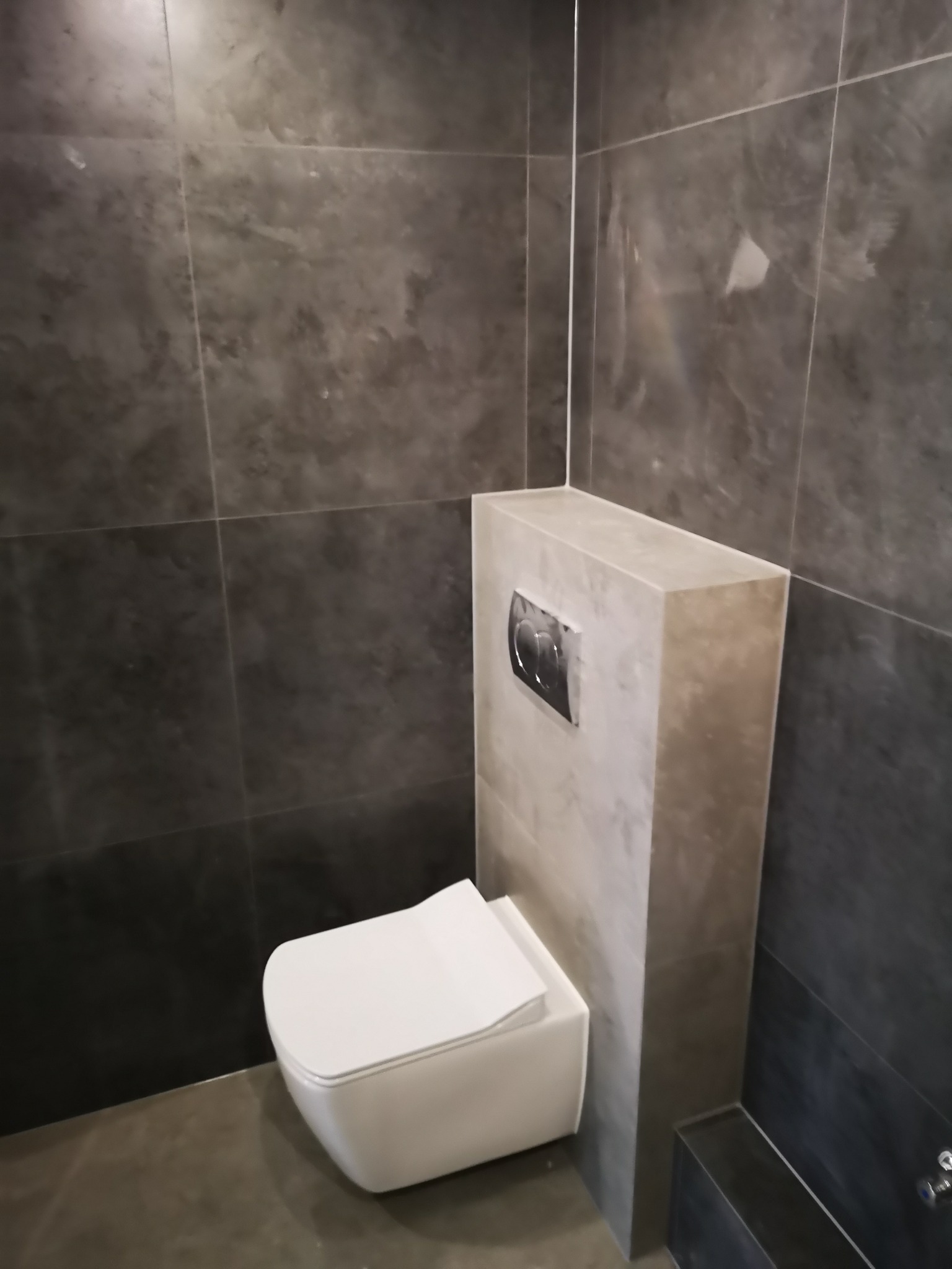 Nowoczesna łazienka z podwieszaną toaletą i szarą glazurą. Minimalistyczny design, widoczny przycisk spłukiwania. Wykończenie w odcieniach szarości.