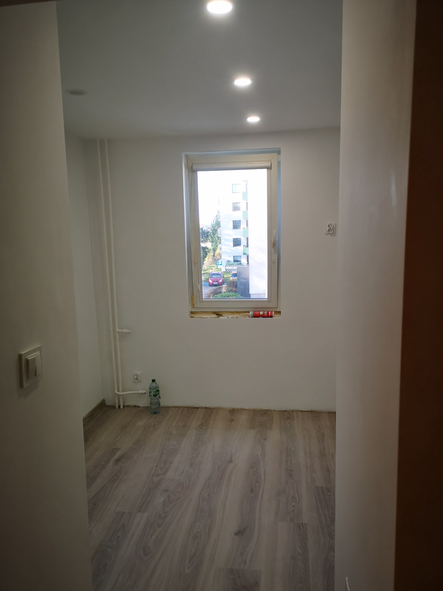 Jasne wnętrze po remoncie: panele podłogowe, białe ściany z grzejnikami i oknem z widokiem na zieleń. Butelka wody przy ścianie. Widoczne ślady po remoncie.