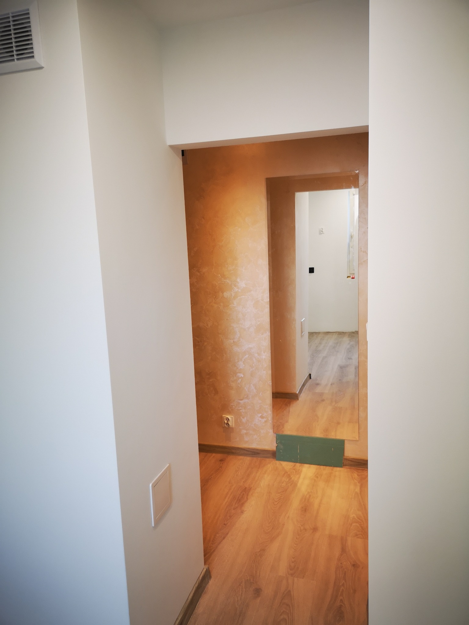 Wykończony korytarz z drewnianą podłogą i ścianami w odcieniach bieli i beżu. Widoczne gniazdko elektryczne i fragment wentylacji. W głębi widoczne kolejne pomieszczenie.