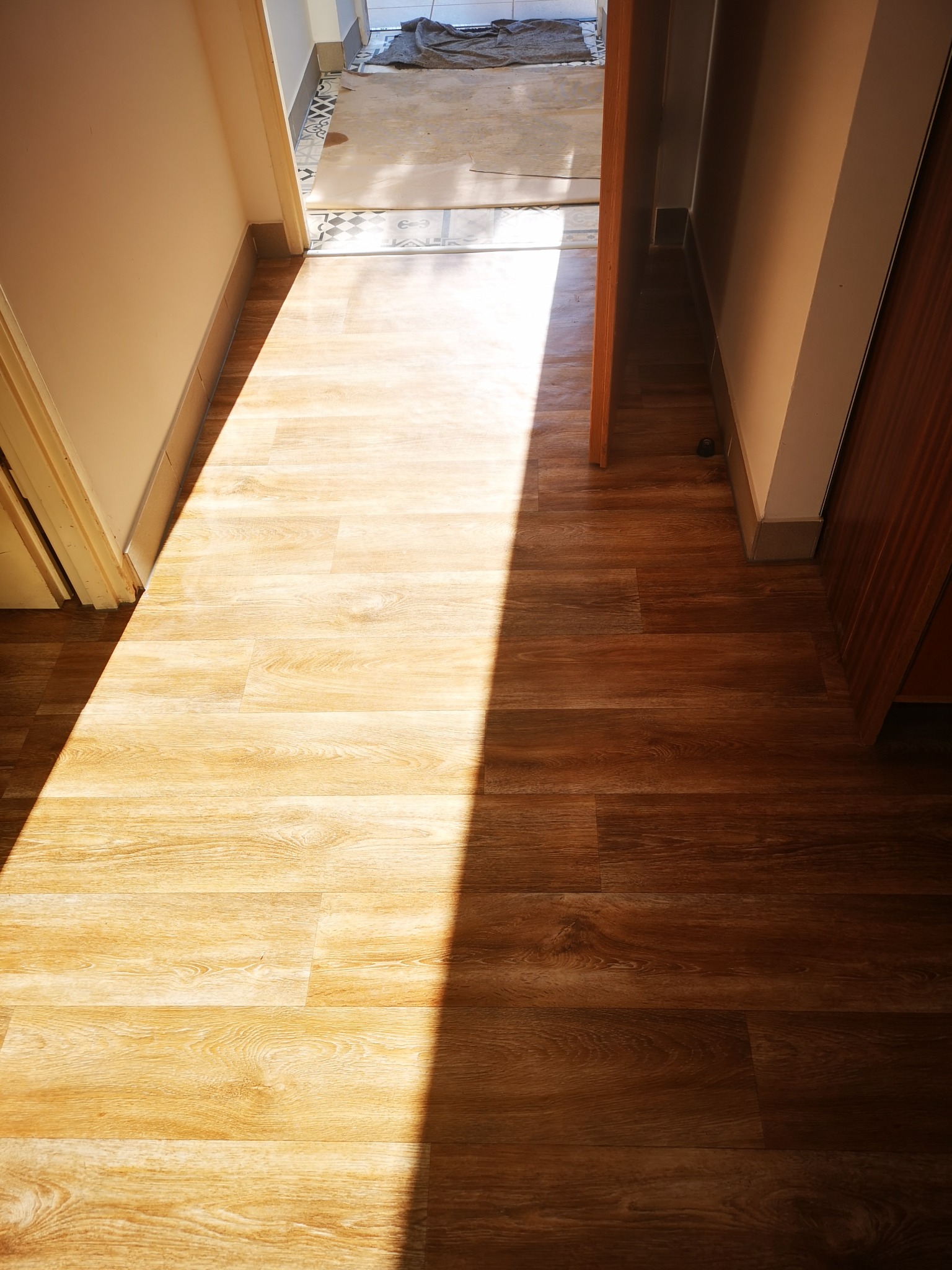 Ułożona podłoga z paneli w korytarzu, z widocznym fragmentem pomieszczenia z płytkami. Wpadające światło podkreśla strukturę drewna.