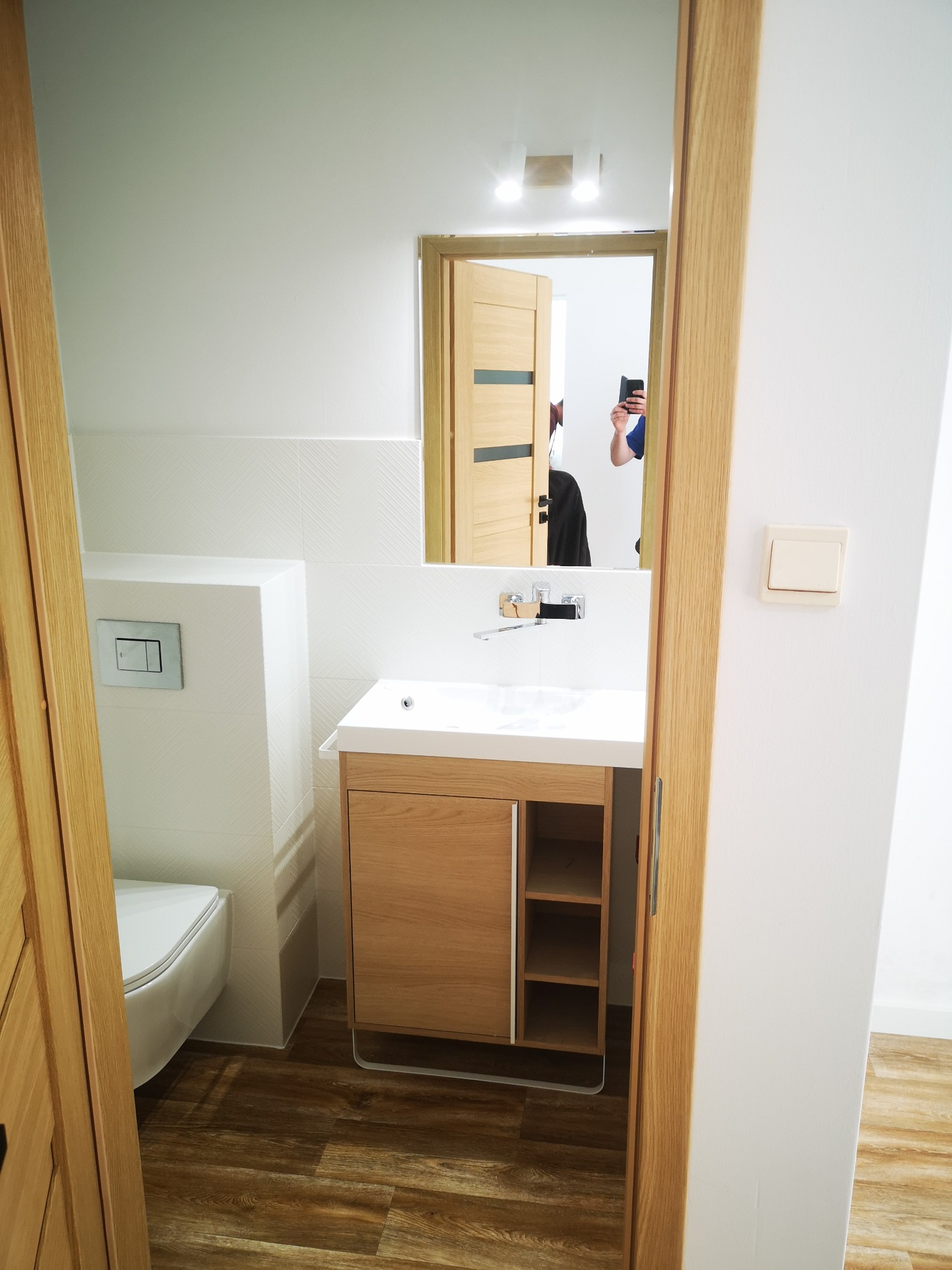 Nowoczesna łazienka po remoncie: białe płytki z subtelnym wzorem, drewniana szafka pod umywalką z otwartymi półkami, podłoga imitująca drewno, widok zza otwartych drzwi.