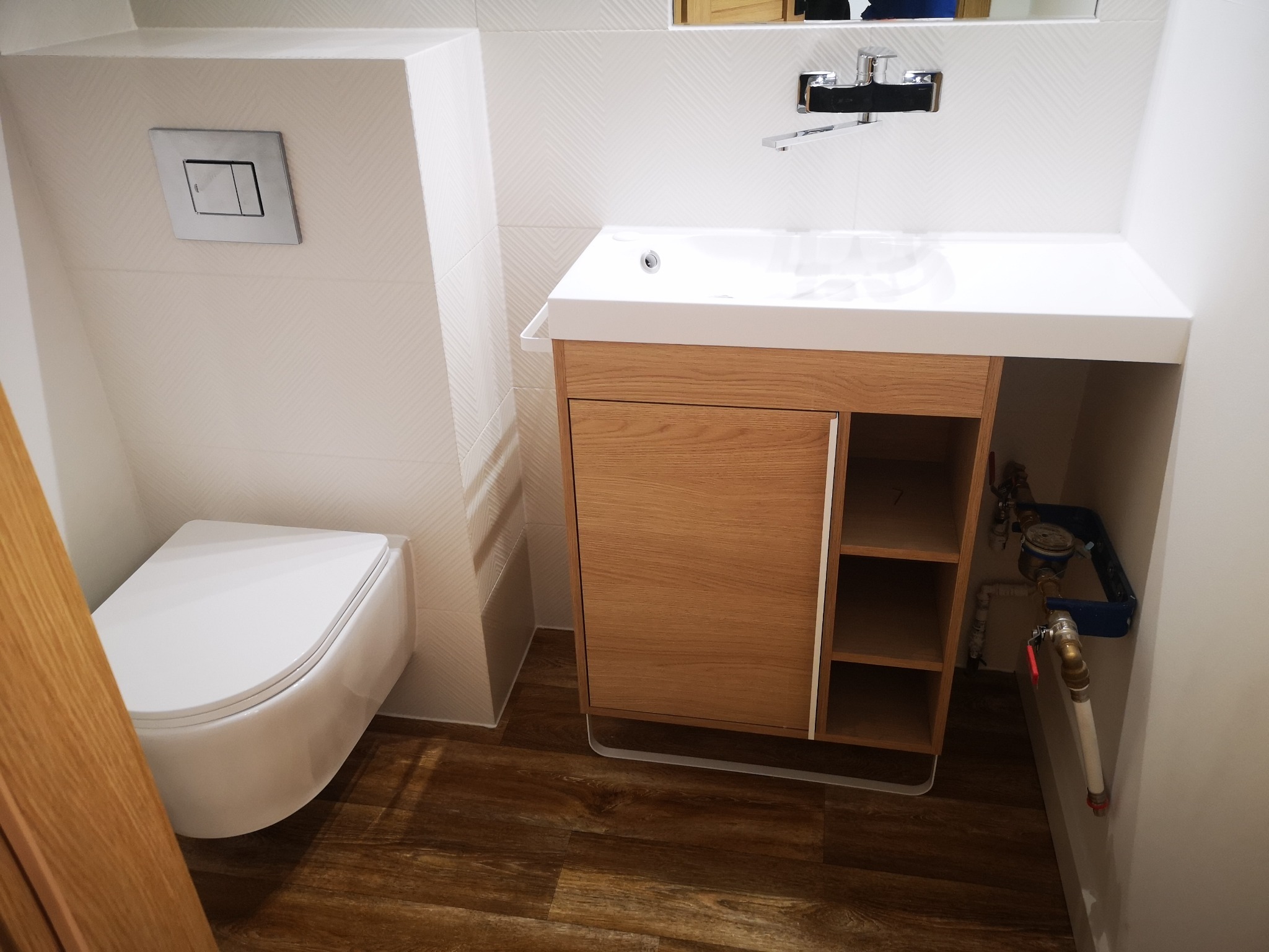 Nowoczesna łazienka po remoncie: podwieszana toaleta, drewniana szafka pod umywalką z blatem, białe płytki z subtelnym wzorem jodełki i drewnopodobna podłoga.