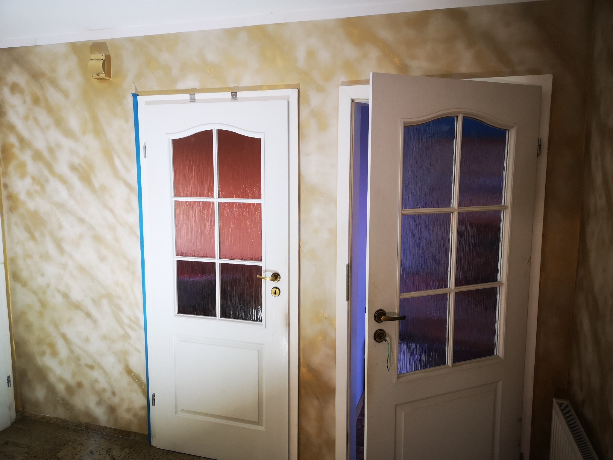 Dwie białe, przeszklone drzwi wewnętrzne po montażu, z widoczną dekoracyjną strukturą ściany w odcieniach beżu i złota. Jedne drzwi otwarte, drugie zamknięte.