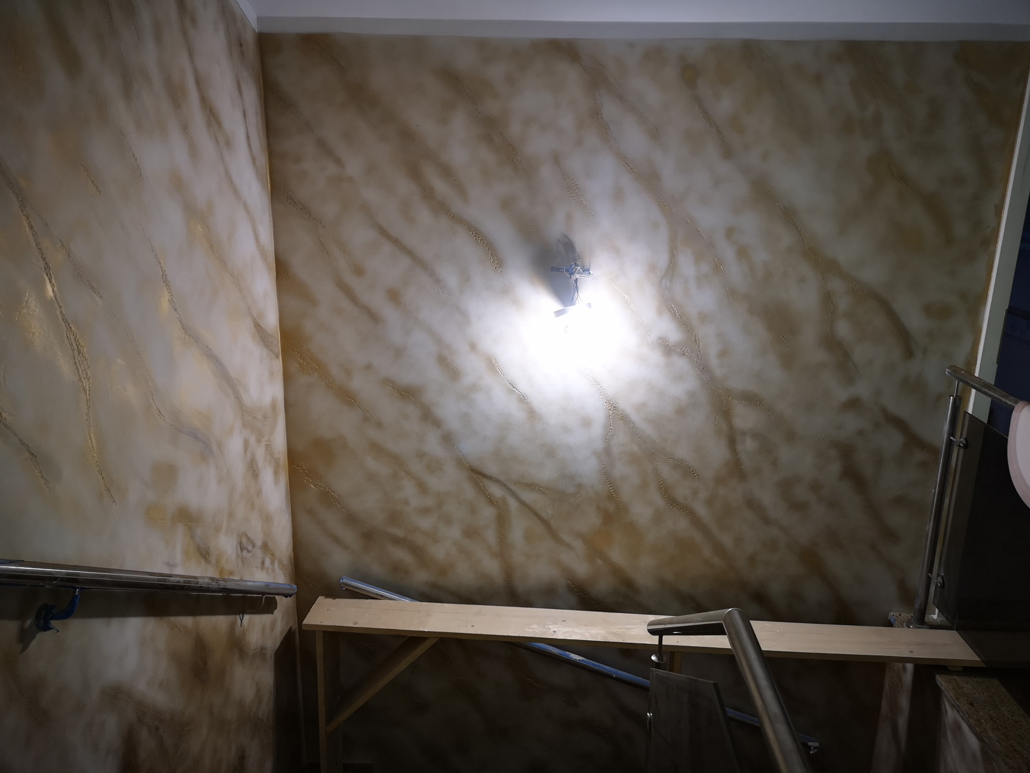 Ściana z efektem dekoracyjnym w odcieniach beżu i złota, oświetlona lampą. Widoczne fragmenty rusztowania i poręczy. Dekoracyjne malowanie ścian.