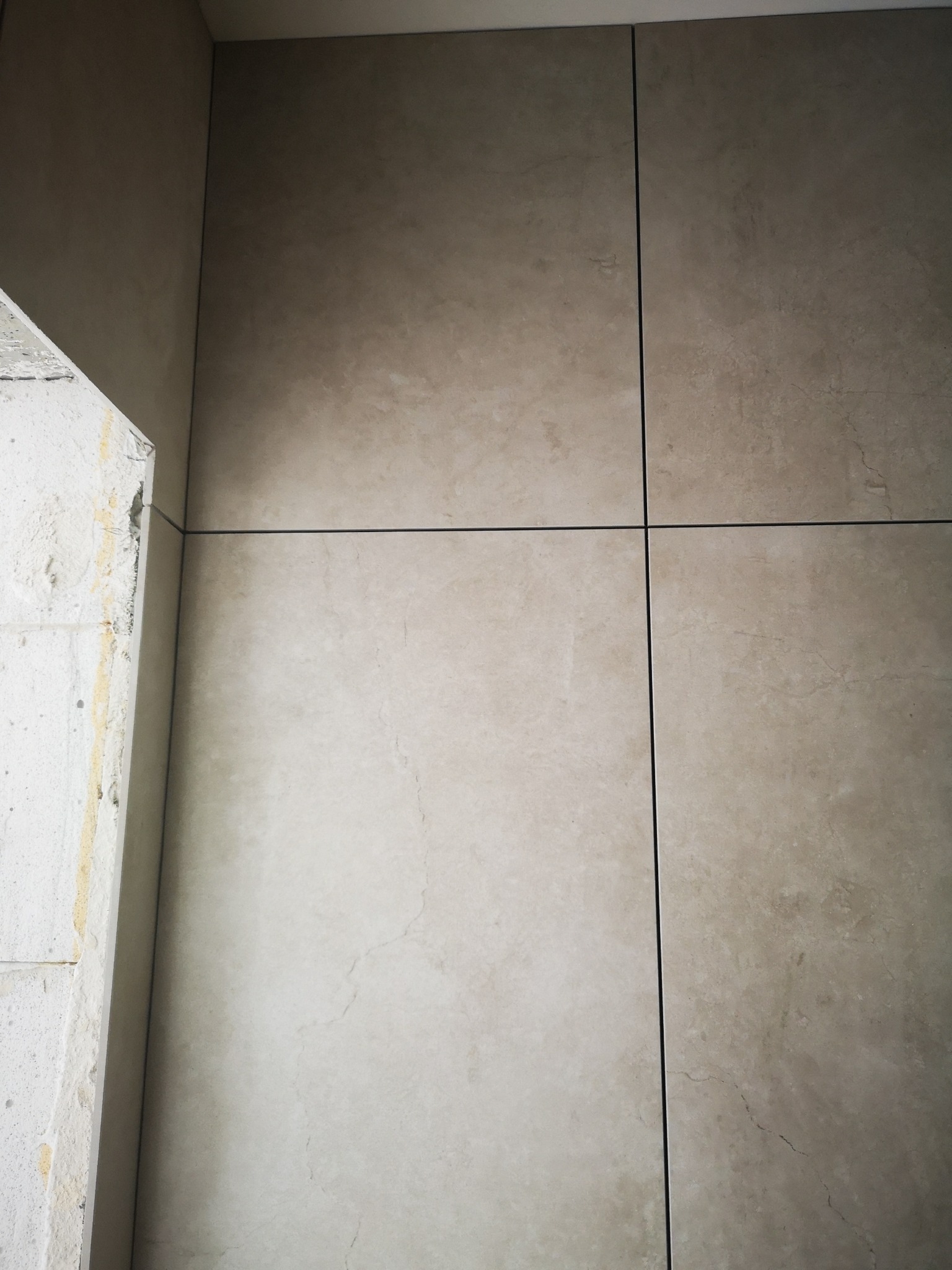 Ściana wyłożona beżowymi płytkami z ciemnymi fugami, widoczny fragment surowej ściany z lewej strony. Ujęcie z bliska, podkreślające fakturę i precyzję ułożenia.