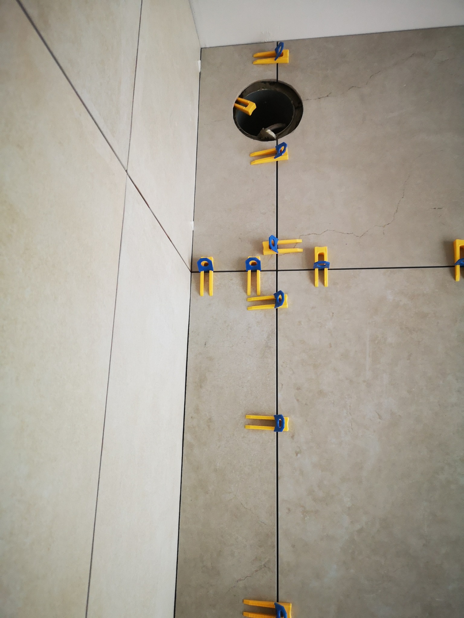 Ściana z beżowych płytek, z żółto-niebieskimi klipsami poziomującymi i czarnymi krzyżykami. Widoczny otwór na rurę. Ujęcie z bliska, ukazujące precyzję układania.