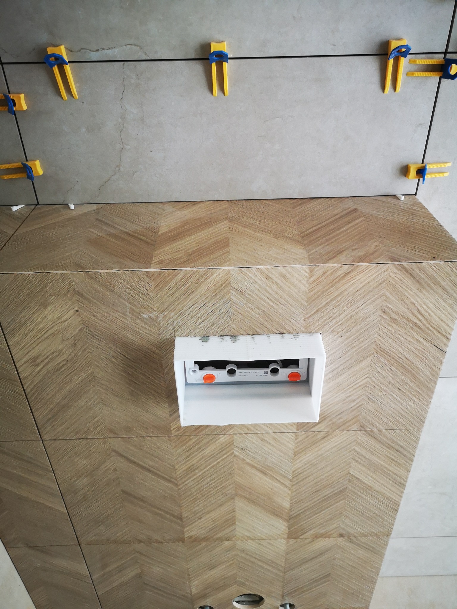 Ułożona glazura w jodełkę na ścianie z widoczną puszką instalacyjną i systemem poziomowania płytek. Detal wykończenia wnętrza łazienki w trakcie prac.