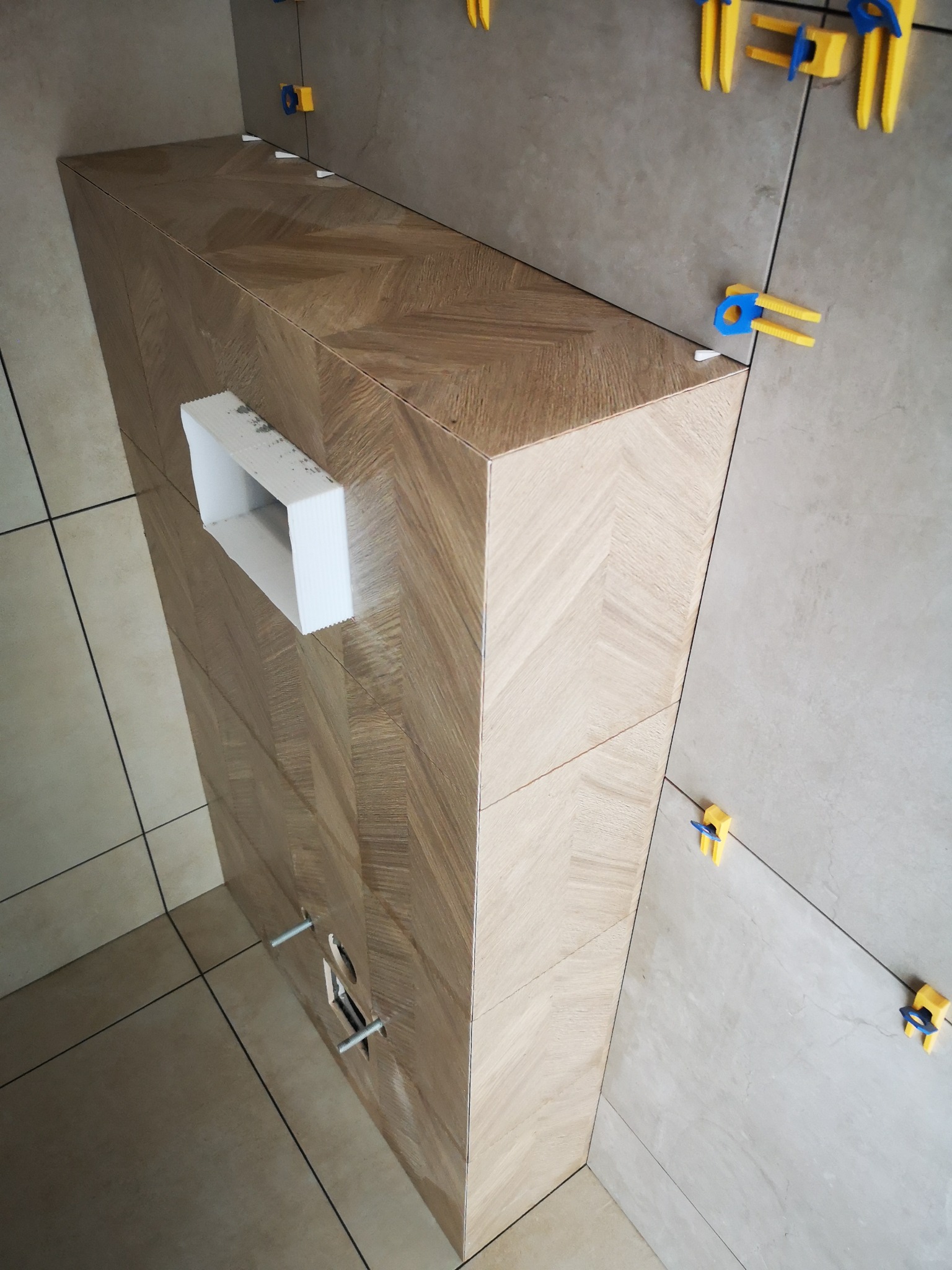 Obudowa stelaża WC wykończona płytkami imitującymi drewno w jodełkę, z widocznymi elementami montażowymi i poziomującymi płytki. Jasne płytki na ścianie i podłodze.