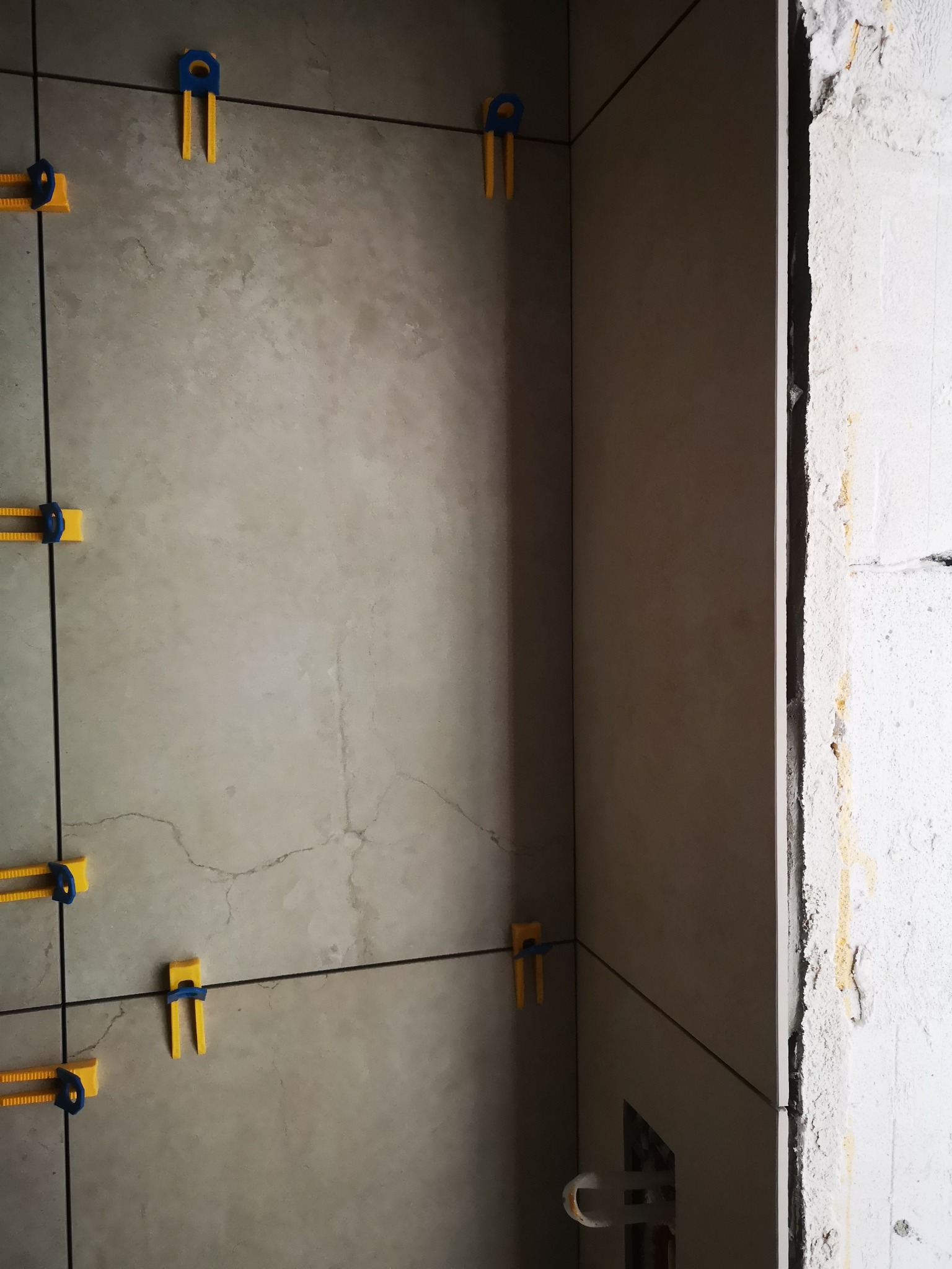 Ściana z szarych płytek w trakcie układania, z widocznymi klipsami poziomującymi w kolorze żółto-niebieskim. Surowa ściana obok, widoczne rury.