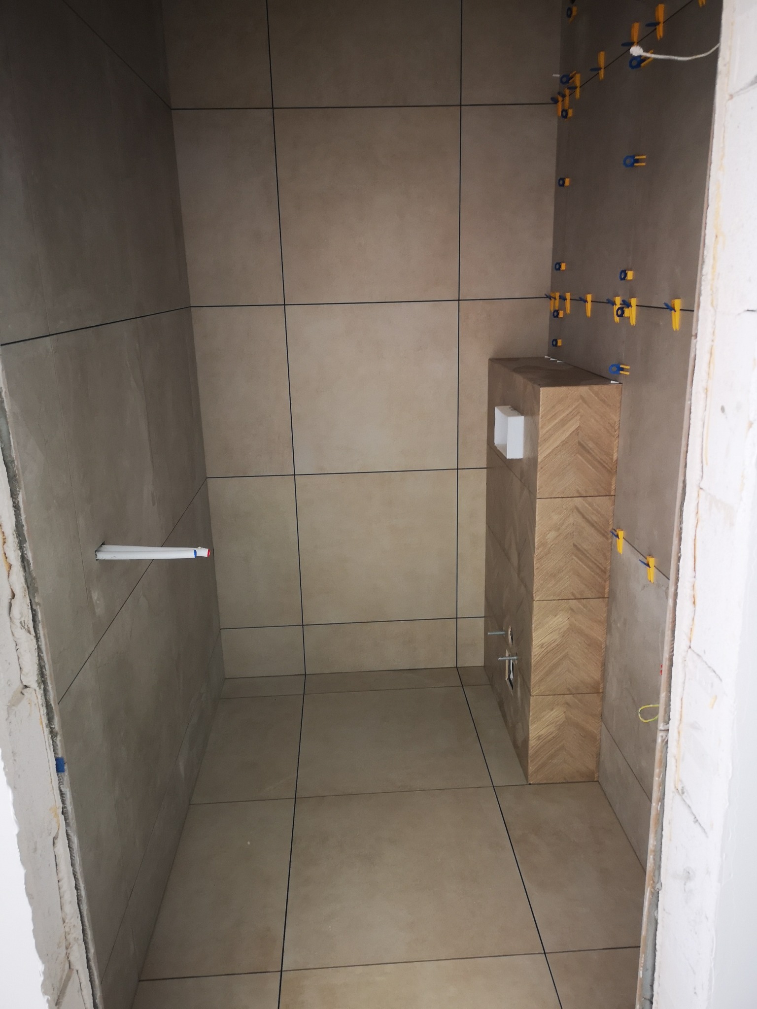 Wnętrze łazienki w trakcie wykańczania: beżowe płytki na ścianach i podłodze, widoczne elementy instalacji i poziomowania płytek. Uwagę przykuwa obudowa WC z wzorem jodełki.