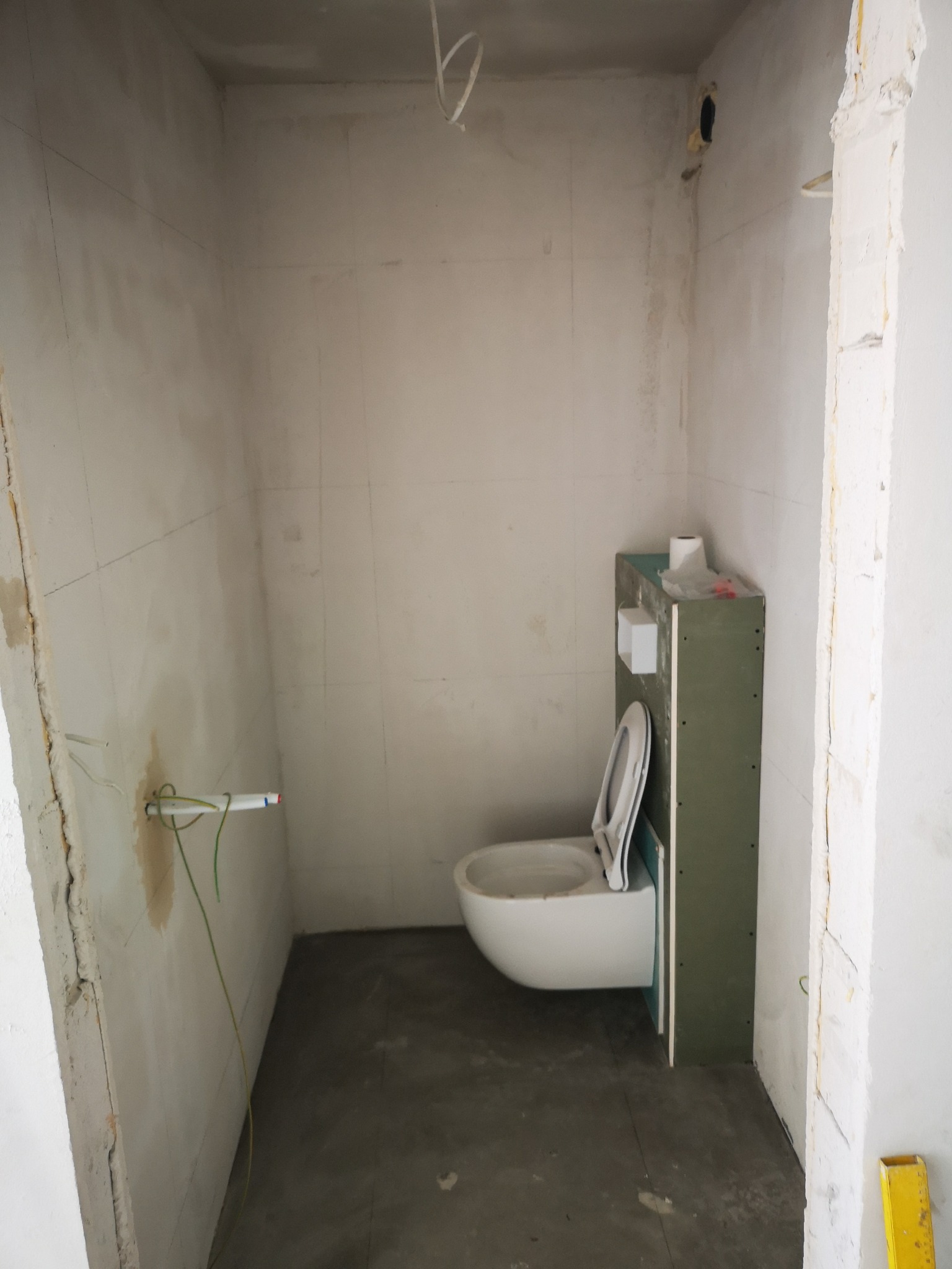 Wnętrze łazienki w trakcie remontu: widoczne przewody, surowe ściany, częściowo zamontowana toaleta podwieszana z obudową z płyty gipsowej. Szary beton na podłodze.