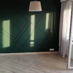 Nowoczesne wnętrze: ciemnozielona ściana z geometrycznym wzorem wykonanym z listew, drewniana podłoga, beżowa lampa wisząca i zasłony w oknie.