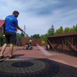 Mężczyzna w niebieskiej koszulce z logo firmy czyści ciśnieniowo brukowaną podjazd z wzorem koła, widoczna myjka ciśnieniowa, brama w tle.