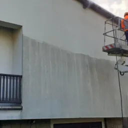 Dwóch pracowników na podnośniku koszowym czyści elewację dwupiętrowego domu, widoczne ślady po czyszczeniu, dom z balkonem.