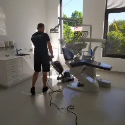 Pracownik w ciemnej koszulce z logo firmy czyści podłogę w gabinecie dentystycznym za pomocą maszyny czyszczącej; widoczne białe meble, okno z widokiem na zieleń i nowoczesny fotel dentystyczny.