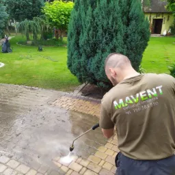 Mężczyzna w koszulce z logo firmy czyści kostkę brukową myjką ciśnieniową na tle zielonego ogrodu z domem.