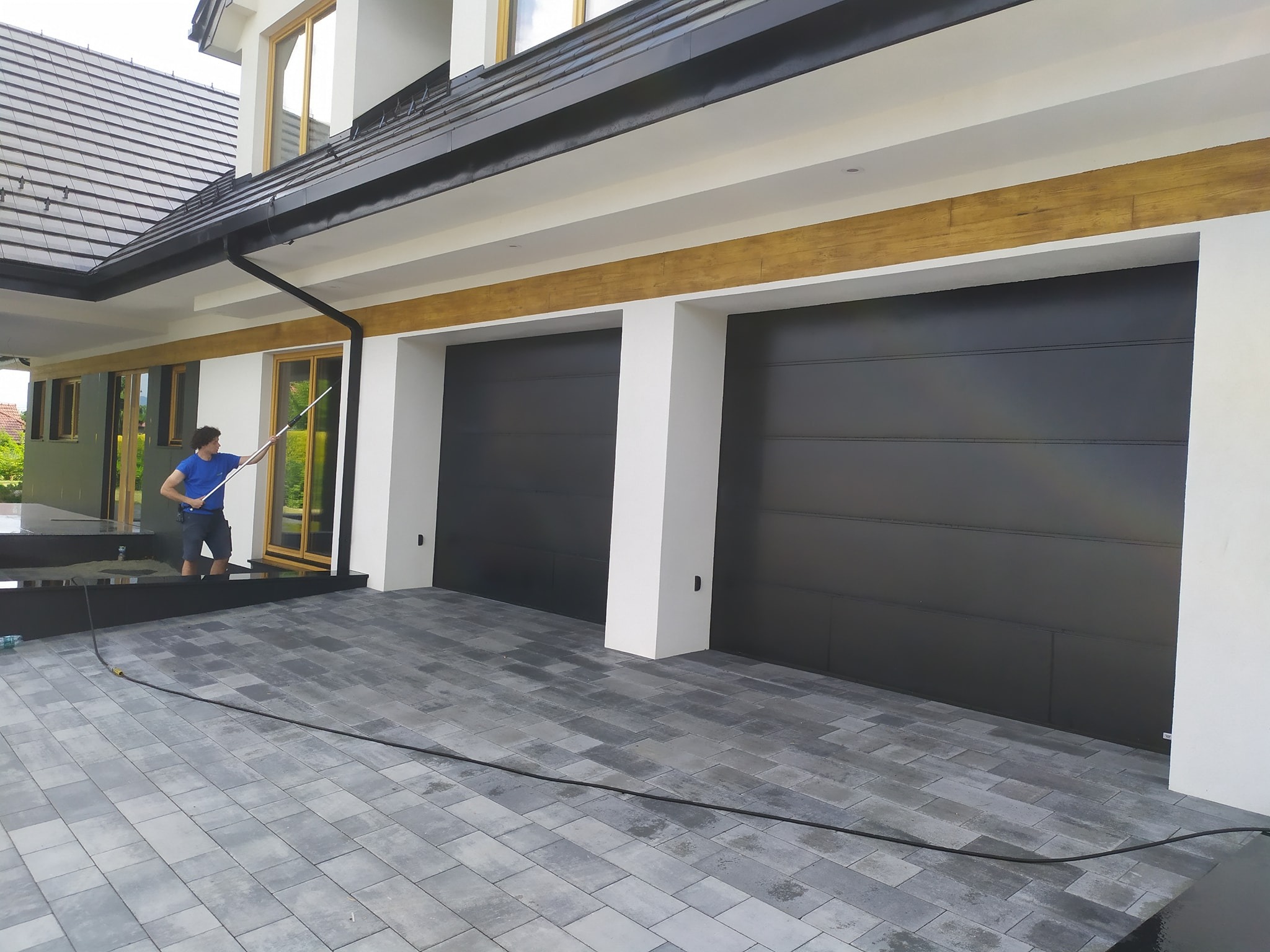 Mężczyzna w niebieskiej koszulce myje elewację nowoczesnego domu z czarnymi bramami garażowymi, używając teleskopowej lancy i węża leżącego na szarej kostce brukowej.