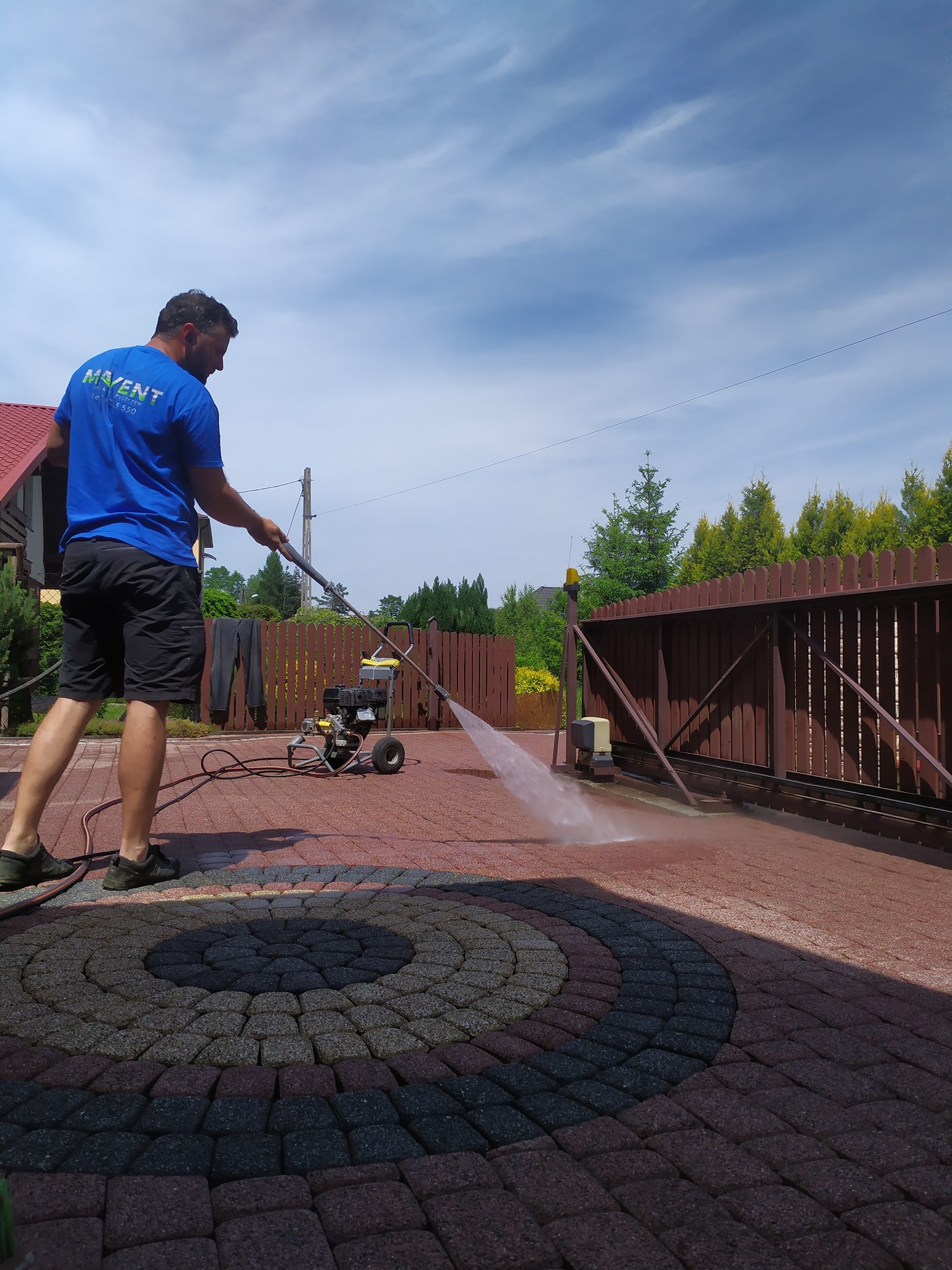 Mężczyzna w niebieskiej koszulce z logo firmy czyści ciśnieniowo brukowaną podjazd z wzorem koła, widoczna myjka ciśnieniowa, brama w tle.