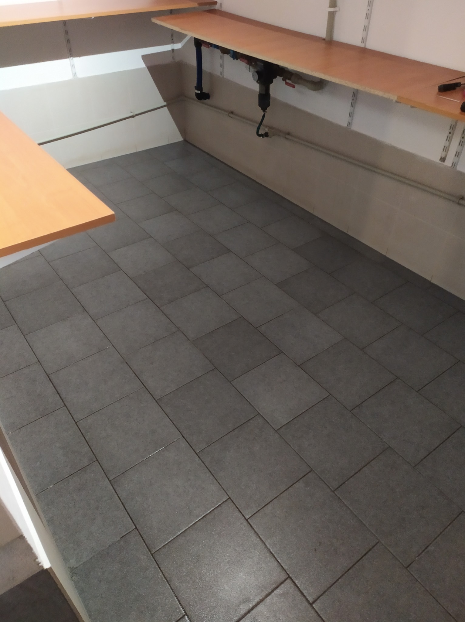 Szare płytki podłogowe po czyszczeniu, widoczne jasne blaty robocze w tle, oświetlenie sufitowe.