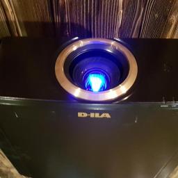 Agra Art Rafał Dogoda - Czarny projektor D-ILA zamontowany na drewnianym suficie z widocznym obiektywem z niebieskim światłem.