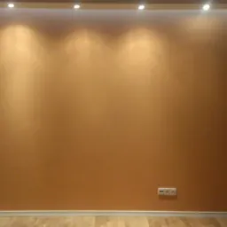 Pomarańczowa ściana w pokoju z drewnianą podłogą, oświetlona przez punktowe światła w suficie, widoczne gniazdka elektryczne i włączniki.