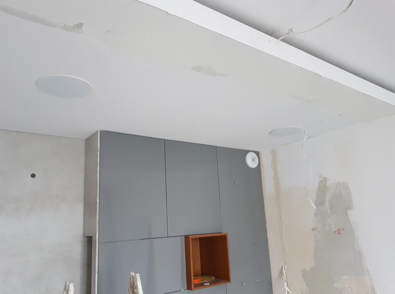 Wnętrze w trakcie prac wykończeniowych: szare szafki w zabudowie, widoczne przewody elektryczne, dwa okrągłe głośniki sufitowe i czujnik dymu na suficie, ściany częściowo otynkowane.