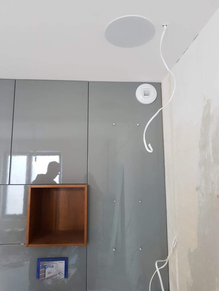 Szara, lakierowana zabudowa ściany z wnęką z jasnego drewna i okrągłym głośnikiem sufitowym; widoczne kable i otwór wentylacyjny w ścianie.