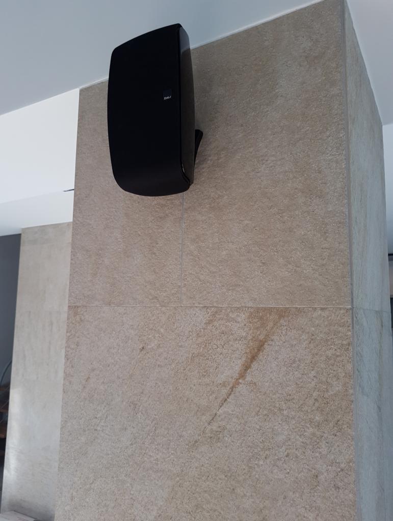 Czarny głośnik marki Dali zamontowany na ścianie wyłożonej płytkami imitującymi kamień w odcieniach beżu i brązu. Widoczny fragment sufitu.