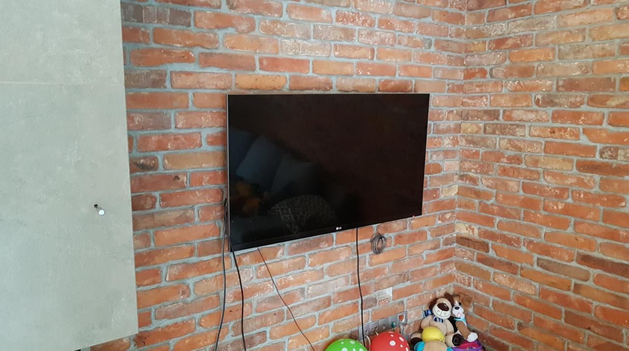 Telewizor LG zawieszony na ceglanej ścianie w rogu pokoju, widoczne kable, obok ściana wykończona płytami imitującymi beton, na podłodze zabawki.