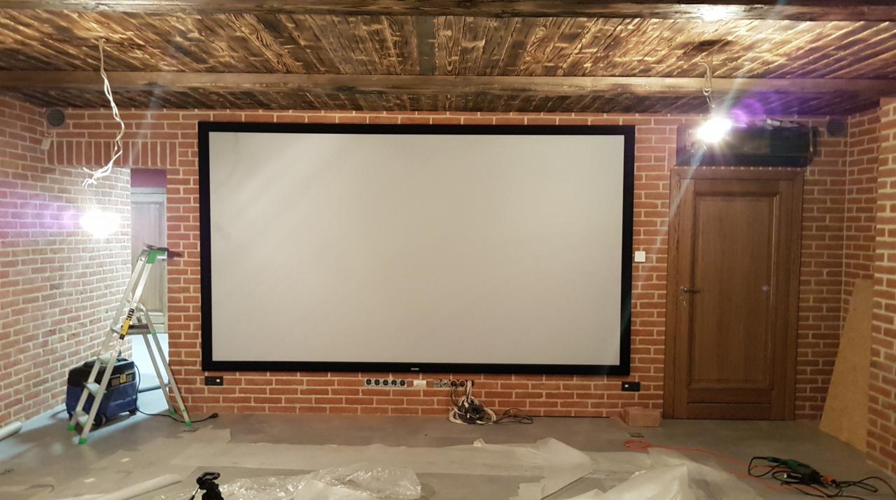 Montaż ekranu projekcyjnego w pomieszczeniu z ceglanymi ścianami i drewnianym sufitem; widoczna drabina, sprzęt elektryczny i częściowo zakryta podłoga.